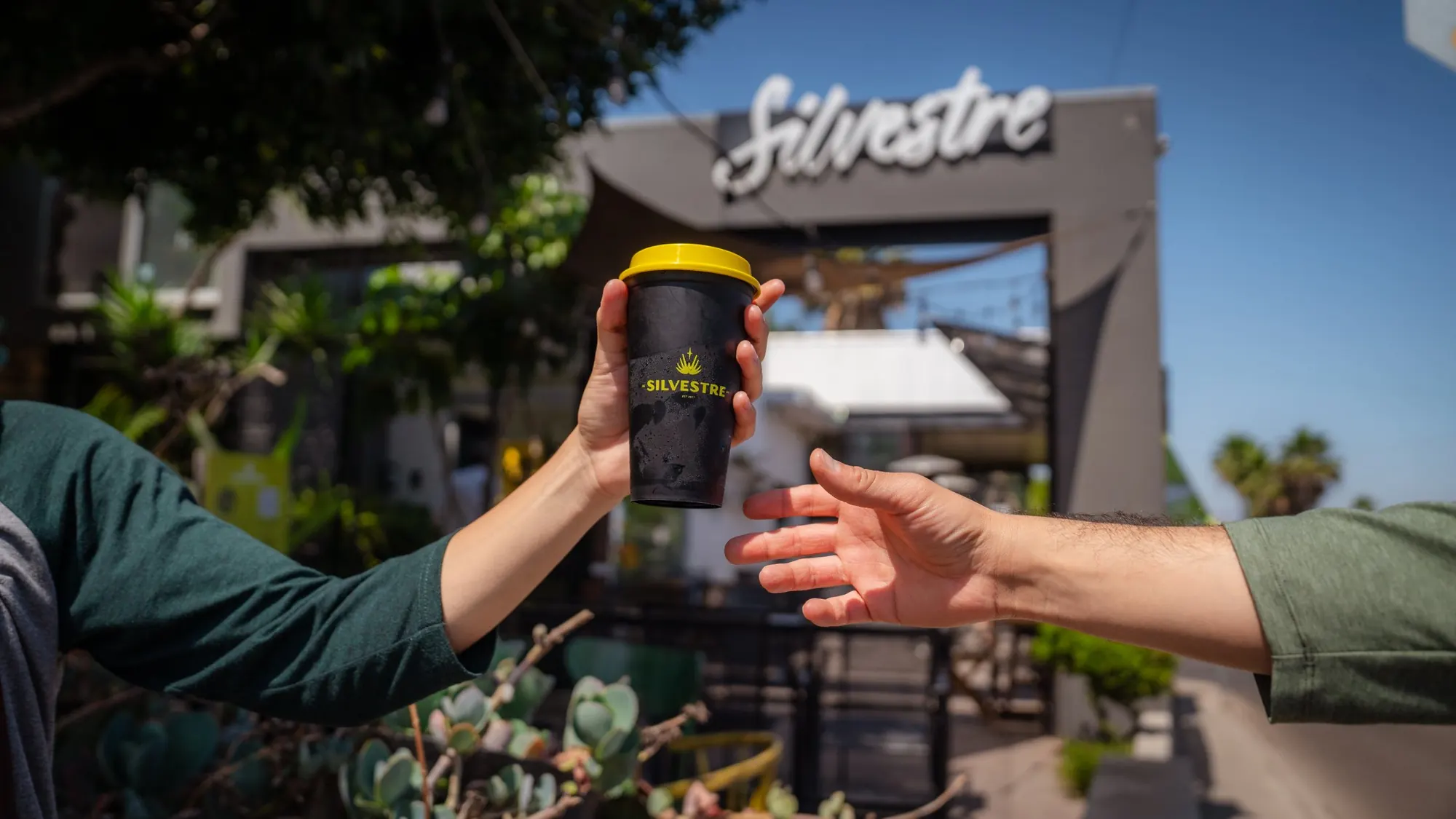 Two hands passing a Silvestre reusable cup in front of the coffee shop